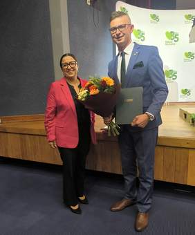 Pokaż zdjęcie: Monika Wac, dyrektor Oddziału Lubelskiego PFRON, i Mateusz Winiarski, dyrektor Oddziału Regionalnego KRUS w Lublinie, pozują do zdjęcia z kwiatami i pamiątkowym grawertonem podczas obchodów 35-lecia KRUS.