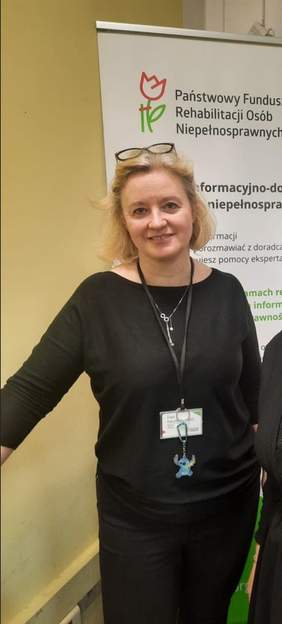 Pokaż zdjęcie: Na zdjęciu stoi kobieta, która się uśmiecha. Jest ubrana w czarną bluzkę. Za nią znajduje się baner z napisem: Państwowy Fundusz Rehabilitacji Osób Niepełnosprawnych. 