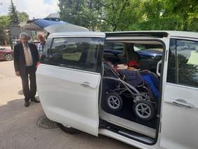 Pokaż zdjęcie: We wnętrzu samochodu znajduje się osoba siedząca na wózku inwalidzkim. Za pojazdem stoi dwóch mężczyzn, którzy oglądają auto. W tle widać zieleń i inne samochody. 
