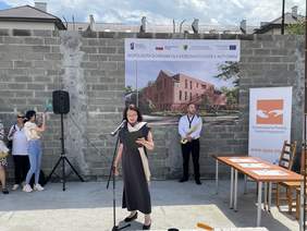 Pokaż zdjęcie: Kobieta w ciemnej sukience przepasana jasnym szalem czyta do mikrofonu teks z kartki, którą trzyma w ręku. Za nią surowy mów na nim plakat z wizualizacją budynku z cegły. Pod murem stoi mężczyzna w białej koszuli z saksofonem.