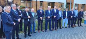 Pokaż zdjęcie: Grupa kilkunastu mężczyzn w eleganckich garniturach stojąca na tle budynku z drewnianymi elementami i oknami. W tle widać ogród z roślinami. Mężczyźni rozmawiają lub pozują do zdjęcia.