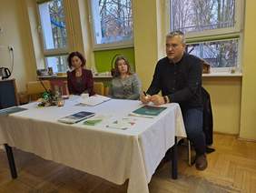 Pokaż zdjęcie: Przy stole prezydialnym, przykrytym białym obrusem siedzą trzy osoby, dwie kobiety i mężczyzna