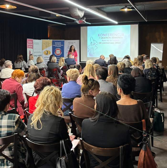 Pokaż zdjęcie: Sala konferencyjna z dużą liczbą osób siedzących tyłem do aparatu, zwróconych w stronę sceny. Na scenie znajduje się kobieta przemawiająca do zgromadzonych. W tle widać biały ekran z prezentacją, na której widnieje napis „KONFERENCJA #JESTEM ZDROWA”. Po lewej stronie ekranu znajdują się kolorowe roll-upy z napisami i logotypami. Ludzie są ubrani elegancko, większość uczestników to kobiety. Pomieszczenie ma nowoczesny wystrój, widoczne są elementy instalacji na suficie.