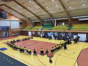 Pokaż zdjęcie: Widok hali sportowej z góry. Osoby poruszające się na wózkach tworzą duży okrąg na środku boiska. W tle znajdują się trybuny, dekoracje z balonów oraz stanowiska rozmieszczone przy ścianie hali.)