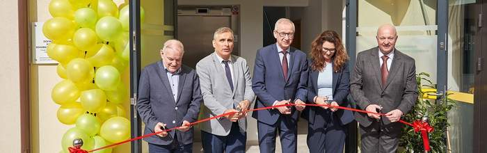Pokaż zdjęcie: Przed wejściem do budynku czterech mężczyzn w garniturach i kobieta w marynarce trzymają w rękach czerwoną wstęgę i nożyczki. Po prawej stronie wejścia mnóstwo balonów spiętych w łuk.