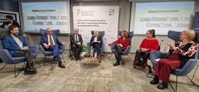 Pokaż zdjęcie: Siedmioro panelistów siedzi na fotelach na środku sali. Moderatorka prowadząca dyskusję siedzi na środku.