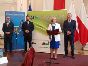 4 osoby stoją w szeregu, trzech mężczyzn i jedna kobieta, kobieta przemawia do mikrofonu, za nimi banery flagi biało-czerwone