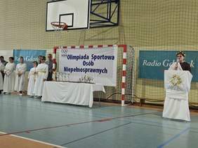 Pokaż zdjęcie: Olimpiada Sportowa Osób Niepełnosprawnych w hali sportowej. Przy bramce i stole z pucharami stoi grupa osób ubranych w białe stroje stylizowane na antyczne. Po prawej osoba przemawia przy mównicy ozdobionej wieńcem laurowym.