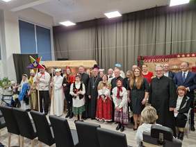 Wykonawcy jasełek i goście stoją razem, mężczyźni i kobiety, grupa kilkunastu osób, większość kolorowo ubrana, z elementami świątecznymi