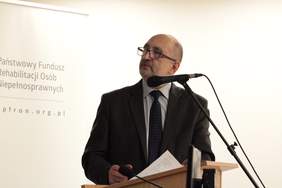 Pokaż zdjęcie: Mężczyzna ubrany w garnitur stoi przy mikrofonie spogląda na ścianę z prezentacją multimedialną
