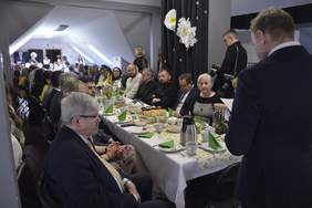 Pokaż zdjęcie: Przy podłużnych stołach siedzą ludzie. Trwa wielkanocne śniadanie. Na stołach leżą świąteczne potrawy, ciasta i dekoracje. 