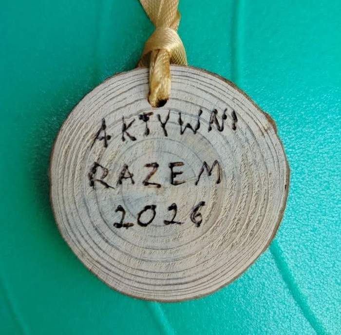 Pokaż zdjęcie: Drewniany medal w kształcie krążka z widocznymi słojami drewna na tle w intensywnym turkusowym kolorze. U góry medalu jest przewleczona żółtawa wstążka. Na środku medalu widnieje odręczny napis wypalony w drewnie: Aktywni Razem 2026.