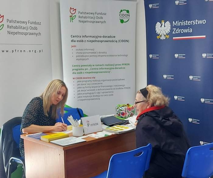 Pokaż zdjęcie: Przy stole siedza dwie kobiety, jedna z nich notuje coś na kartce. Z tyłu za stolikiem stoją banery Ministerstwa Zdrowia, CIDON oraz PFRON