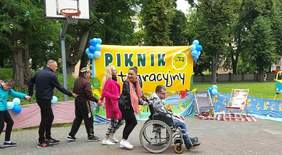 Pokaż zdjęcie: Na boisku wyłożonym kostką brukową znajduje się mężczyzna na wózku, a za nim pięć innych osób trzymających się za ręce. W tle baner przyozdobiony balonami z napisem: „Piknik integracyjny”, leżaki, kosz do gry w koszykówkę oraz drzewa liściaste.