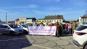 Pokaż zdjęcie: Na miejskim placu w Poddębicach stoi duża grupa osób. Trzymają przed sobą transparent z napisem: Marsz świadomości i akceptacji autyzmu. W tle widać domy i samochody.  