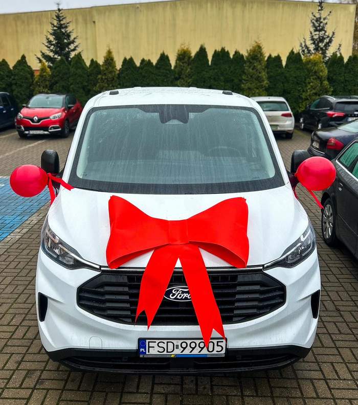 Pokaż zdjęcie: Biały bus widoczny od strony maski, który jest owinięty w czerwoną kokardę i baloniki