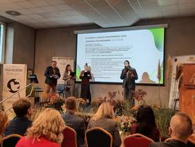 Pokaż zdjęcie: Na scenie stoją cztery osoby. Jedna z pań przemawia do mikrofonu. Na tablicy napisany jest temat spotkania: III Forum z okazji Światowego Dnia Zdrowia Psychicznego „Ptasi Móżdżek” – Łódź, 10 października 2025.