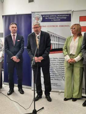 Pokaż zdjęcie: Do mikrofonu przemawia mężczyzna ubrany w garnitur. Po jego bokach stoją: kobieta i drugi mężczyzna. W tle widać baner Powiatowego Centrum Zdrowia w Opocznie. 