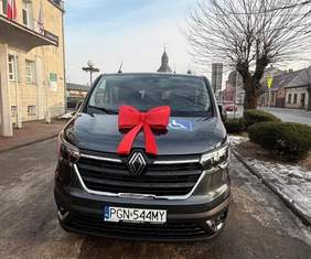 Pokaż zdjęcie: Samochód w kolorze czarnym stoi przodem do zdjęcia. Na masce samochodu ozdobiony jest dużą czerwoną kokardą. W tle budynek po lewej stronie, a po prawej stronie drzewo bez liści, zabudowa miejska, a na ziemi resztki śniegu. 