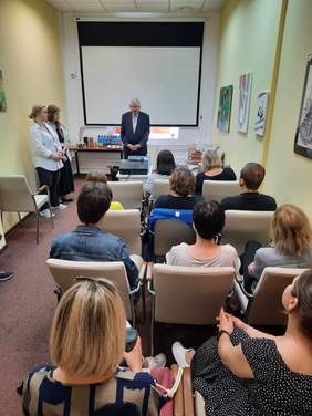 Pokaż zdjęcie: W sali w rzędach siedzą na krzesłach ludzie. Patrzą się na mężczyznę, który stoi przed nimi i przemawia. Obok prowadzącego stoją dwie kobiety. 