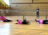 Trzech zawodników w różowych koszulkach leży na parkiecie przed bramką podczas meczu goalball, próbując obronić dostęp do bramki. Zawodnicy mają zasłonięte oczy.