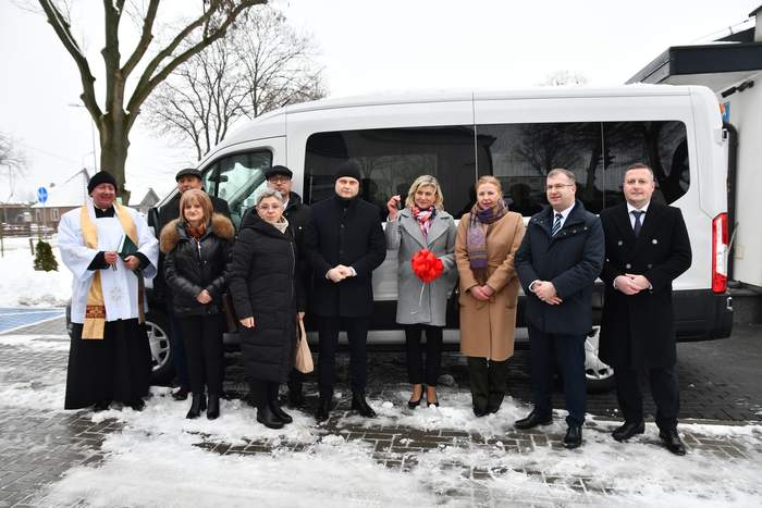 Pokaż zdjęcie: Na tle białego busa pozują uczestnicy uroczystości przekazania kluczyków. Łącznie 10 osób.