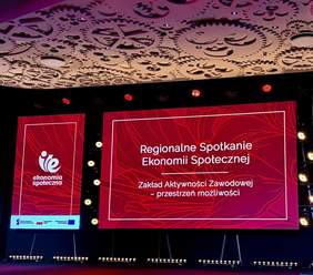 Pokaż zdjęcie: Scena konferencyjna z dużymi czerwonymi ekranami LED prezentującymi napis „Regionalne Spotkanie Ekonomii Społecznej. Zakład Aktywności Zawodowej – przestrzeń możliwości”, oświetlenie sceniczne i dekoracyjny sufit nad sceną.