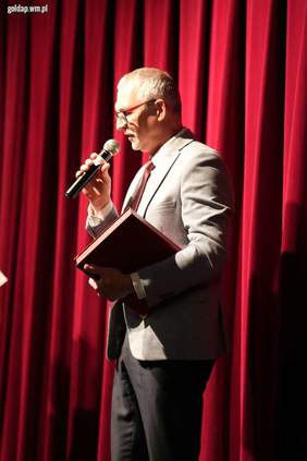 Pokaż zdjęcie: Mężczyzna w garniturze z mikrofonem w dłoni przemawia na scenie na tle czerwonej kurtyny. Zdjęcie wykonane w trakcie oficjalnego wydarzenia.