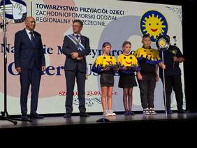 Pokaż zdjęcie: Pięć osób stoi na scenie przed dużym ekranem podczas uroczystości. Po lewej stronie stoi dwóch dorosłych mężczyzn w garniturach. Mężczyzna po prawej z nich ma na sobie szarfę z medalami. Obok nich stoi dwoje dzieci i jeden nastolatek ubrani w czarne spódnice/spodnie i żółte pelerynki ozdobione uśmiechniętymi buźkami z logo Towarzystwa Przyjaciół Dzieci (TPD). Dwie dziewczynki trzymają w rękach talerzyki z ciemnymi przedmiotami (prawdopodobnie pamiątkowymi monetami lub medalami). Najstarszy z grupy młodzieży stoi z rękami skrzyżowanymi z przodu. Po prawej stronie, za młodzieżą, stoi kolejny dorosły mężczyzna, który trzyma coś, co wygląda na ceremonialne koło lub tarczę na długim drążku, ozdobione czerwoną różą. W tle widoczny jest ten sam ekran co na zdjęciu 1, z częściowo widocznym logo TPD oraz tekstem "TOWARZYSTWO PRZYJACIÓŁ DZIECI ZACHODNIOPOMORSKI ODDZIAŁ REGIONALNY W SZCZECINIE".