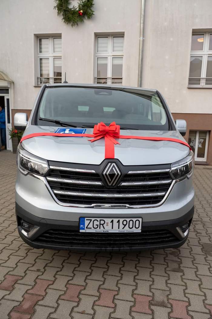 Pokaż zdjęcie: Srebrny van marki Renault stoi zaparkowany przed jasnym budynkiem. Na masce samochodu zawiązana jest duża, czerwona kokarda prezentowa. Na tablicy rejestracyjnej widnieje polski numer rejestracyjny (ZGR 11900). Z przodu na masce, pod kokardą, widoczny jest niebieski znak osoby niepełnosprawnej. Nad samochodem, na fasadzie budynku, widać wieniec świąteczny.