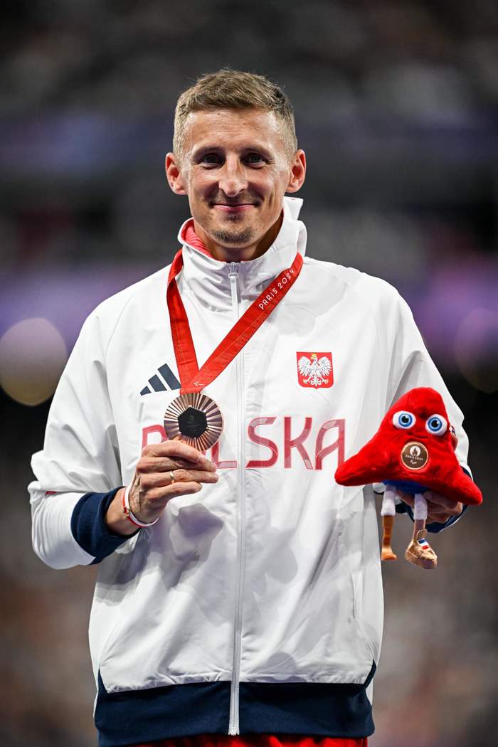 Pokaż zdjęcie: Mężczyna stoi na podium, jest uśmiechnięty, ubrany w sportowy dres, na szyi ma brązowym medal