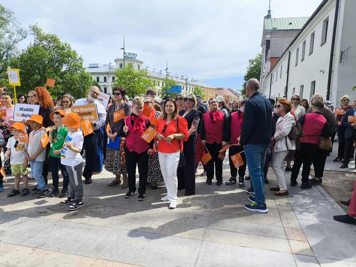 Pokaż zdjęcie: Grupa uczestników pomarańczowego marszu Dnia Godności Osób z Niepełnosprawnością Intelektualną w Lublinie. Dzieci, dorośli i opiekunowie stoją na ulicy, trzymając pomarańczowe chorągiewki i plakaty.