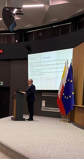 Pokaż zdjęcie: Mężczyzna stojący za mównicą wygłasza prezentację, w tle program konferencji na ekranie.