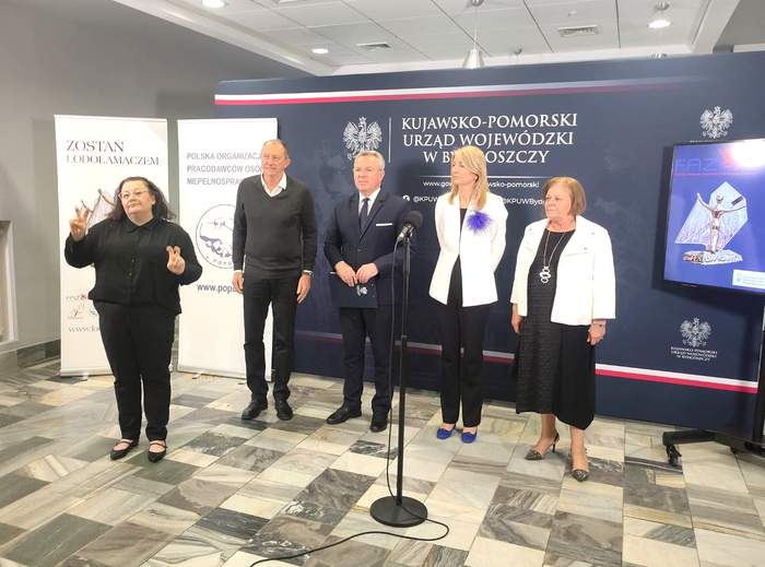 W holu Urzędu Wojewódzkiego stoją od lewej: tłumaczka języka migowego, Pracodawca Osób z Niepełnosprawnościami Holkap sp. z o.o, Wicewojewoda Kujawsko-Pomorski, Dyrektor Generalna Polskiej Organizacji Pracodawców Osób Niepełnosprawnych oraz Dyrektor Kujawsko-Pomorskiego Oddziału PFRON - Aleksandra Gierej. W tle widać banery organizatoró wydarzenia.