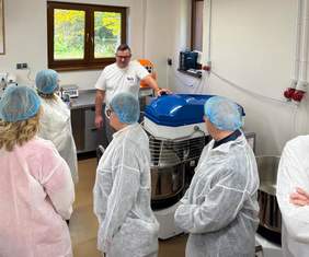 Mężczyzna prezentuje uczestnikom urządzenie wykorzystywane w procesie przetwarzania produktów. Uczestnicy spotkania mają na sobie białe fartuchy ochronne i niebieskie czepki.