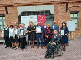 Grupa 13 osób, w tym 1 osoba z prawej strony na wózku inwalidzkim, kobiety i mężczyźni, ubrani elegancko, stoją na tle ściany z cegły, pozują do zdjęcia 