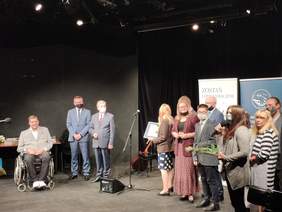Pokaż zdjęcie: Na ciemnej teatralnej scenie stoi grupa nagrodzonych osób
