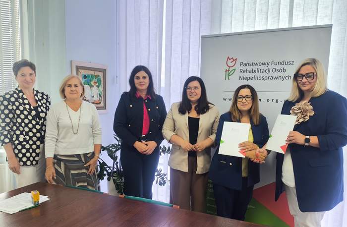 Pokaż zdjęcie: Przy stole konferencyjnym sześć osób pozuje do zdjęcia podczas podpisania umów z PFRON. Dwie osoby trzymają dokumenty, w tle widoczny jest roll-up Funduszu.