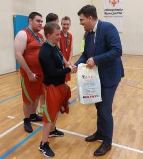 Pokaż zdjęcie: Mężczyzna podaje dłoń zawodnikowi ubranemu w sportowe spodenki i bluzkę, w drugiej ręce trzyma reklamówki, za nimi 3 osoby w strojach sportowych
