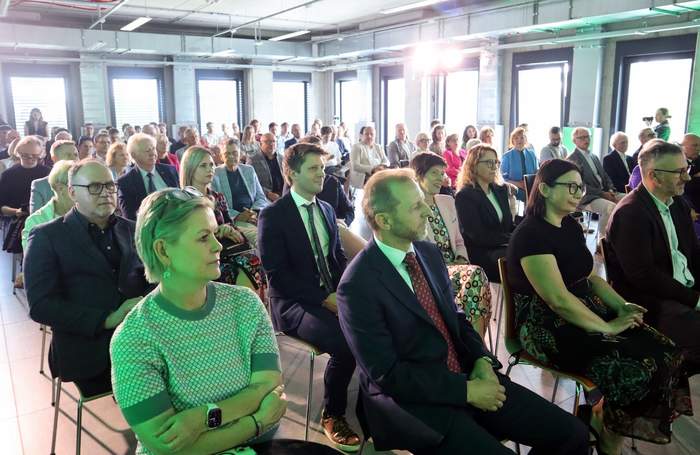 Pokaż zdjęcie: Publiczność siedząca w rzędach w przestronnej sali konferencyjnej z wysokim sufitem podczas finału ogólnopolskiego konkursu architektonicznego. Na pierwszym planie od lewej siedzą: kobieta w jasnozielonej bluzce w kratkę, mężczyzna w ciemnym garniturze z czerwonym krawatem, kobieta w czarnej bluzce oraz mężczyzna w garniturze bez krawata. Wśród zaproszonych gości jest Prezes Zarządu PFRON Małgorzata Lorek oraz dyrektor Oddziału Wielkopolskiego PFRON Anna Skupień. Pomieszczenie oświetla wpadające przez duże okna dzienne światło o lekkim, zielonkawym odcieniu.