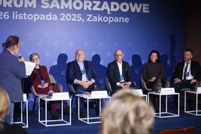 Pokaż zdjęcie: Panel dyskusyjny podczas wydarzenia „Forum Samorządowe” – na scenie siedzi pięcioro prelegentów, obok nich moderator z mikrofonem; w tle widoczny napis „26 listopada 2025, Zakopane”, na pierwszym planie publiczność.