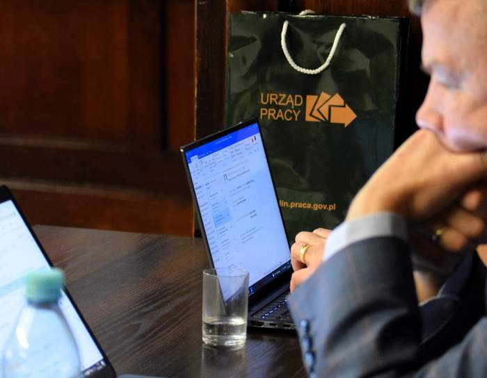 Pokaż zdjęcie: Przy laptopie siedzi mężczyzna wpatrzony w monitor, , mężczyzna ręką zasłania usta.
