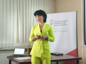 Pokaż zdjęcie: Kobieta brunetka w limonkowym garniturze prowadzi wykład.