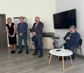 Pokaż zdjęcie: (Po prawej stronie mężczyzna siedzący przed laptopem, po lewej stoi kobieta i dwóch mężczyzn. Za nimi komoda, tablica oraz telewizor. Znajdują się w pomieszczeniu z jasnymi ścianami.