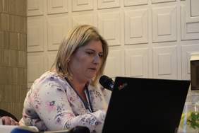 Pokaż zdjęcie: Blondynka z włosami do ramion siedzi przy stole konferencyjnym i mówi do mikrofonu