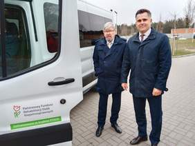 Pokaż zdjęcie: Obok samochodu stoi dwóch mężczyzn, którzy są bardzo elegancko ubrani. Na pojeździe naklejona jest naklejka Państwowego Funduszu Rehabilitacji Osób Niepełnosprawnych. 
