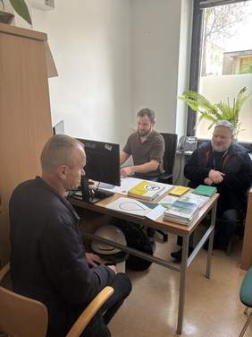 Pokaż zdjęcie: Przy biurku siedzi mężczyzna rozmawiający z dwoma pracownikami podczas dyżuru eksperckiego PFRON. Na stole znajdują się materiały informacyjne i komputer. Spotkanie odbywa się w jasnym pomieszczeniu biurowym z dużym oknem i rośliną w tle.