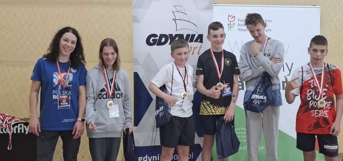 Pokaż zdjęcie: Sześć uśmiechniętych osób, w tym dzieci z medalami i opiekunka, pozuje na tle banerów „Gdynia Sport” i „PFRON”. Uczestnicy mają sportowe stroje i torby z nagrodami, stoją w hali sportowej.