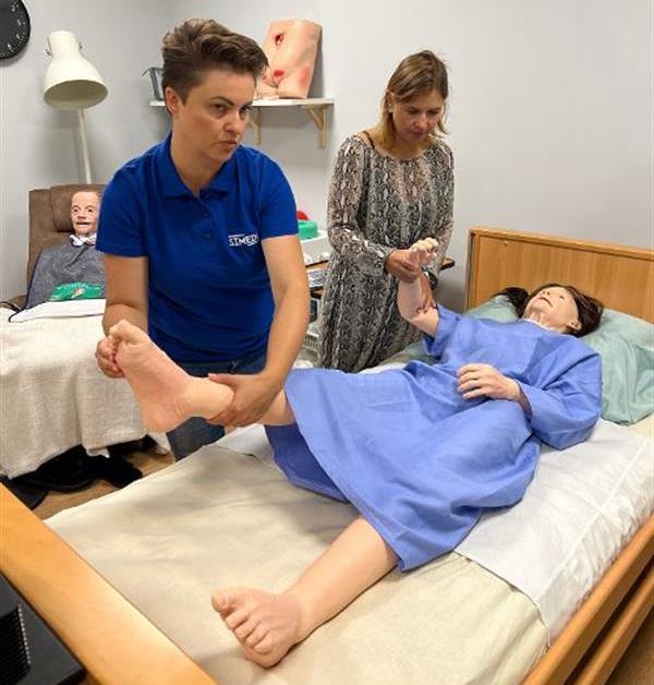 Pokaż zdjęcie: Dwie osoby ćwiczą techniki pielęgnacyjne na realistycznym manekinie leżącym na łóżku. Jedna z nich trzyma nogę manekina, druga podtrzymuje rękę. W tle widać wyposażenie sali szkoleniowej, w tym drugi manekin na łóżku oraz modele anatomiczne.