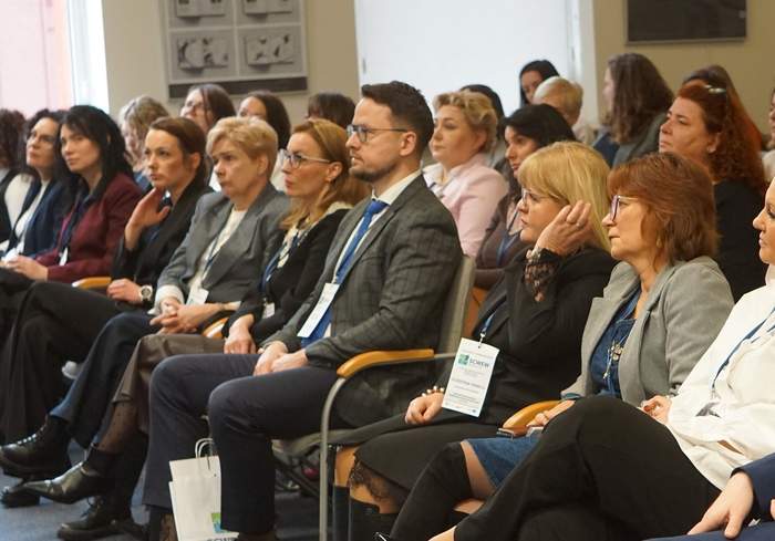Pokaż zdjęcie: Ujęcie przedstawia pierwszy rząd uczestników konferencji siedzących na szarych krzesłach z drewnianymi podłokietnikami. Wśród nich znajduje się mężczyzna w ciemnoniebieskim garniturze oraz kobieta w białej bluzce z falbaną. Większość osób patrzy w prawą stronę (z perspektywy widza), śledząc prezentację lub wystąpienie. Pod krzesłami widoczne są białe torby konferencyjne z logo „SCWEW”. W tle widać pozostałych uczestników oraz fragment okna, przez które wpada dzienne światło.